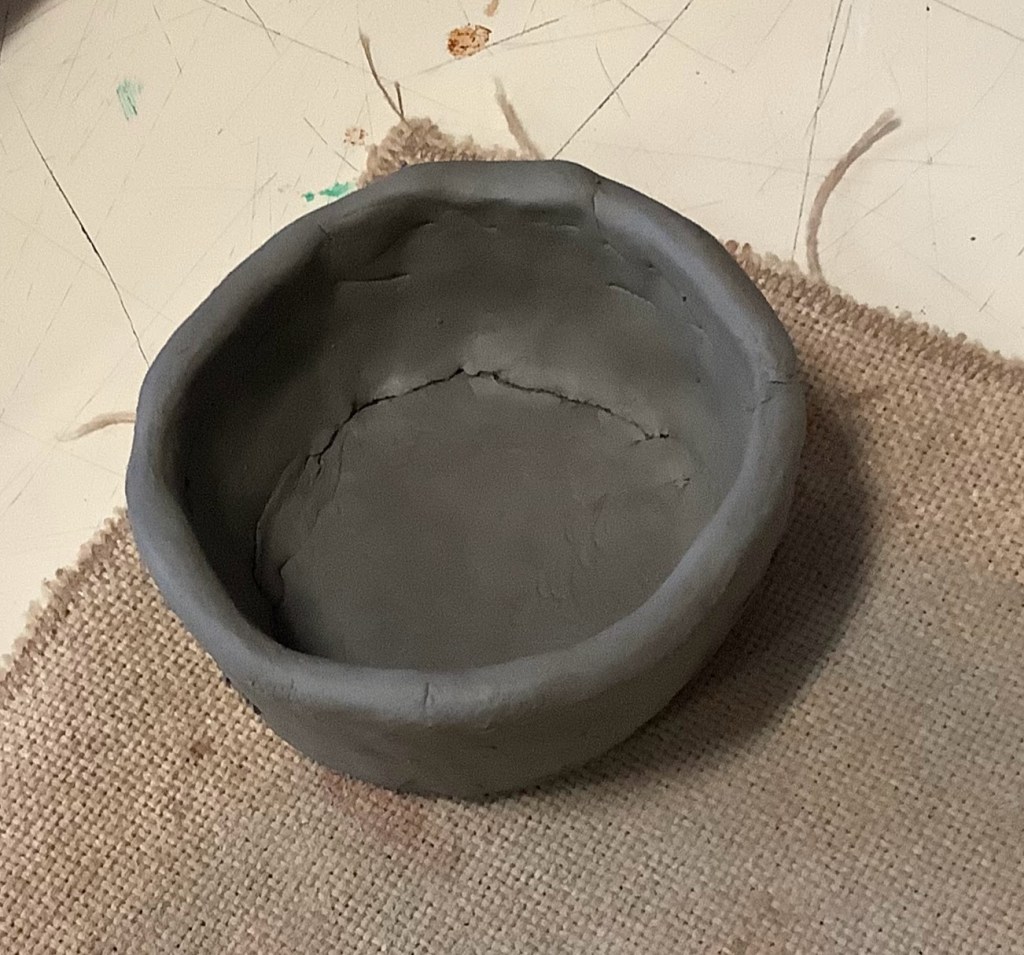 A simple clay bowl or dish in grey clay, resting on a burlap surface.