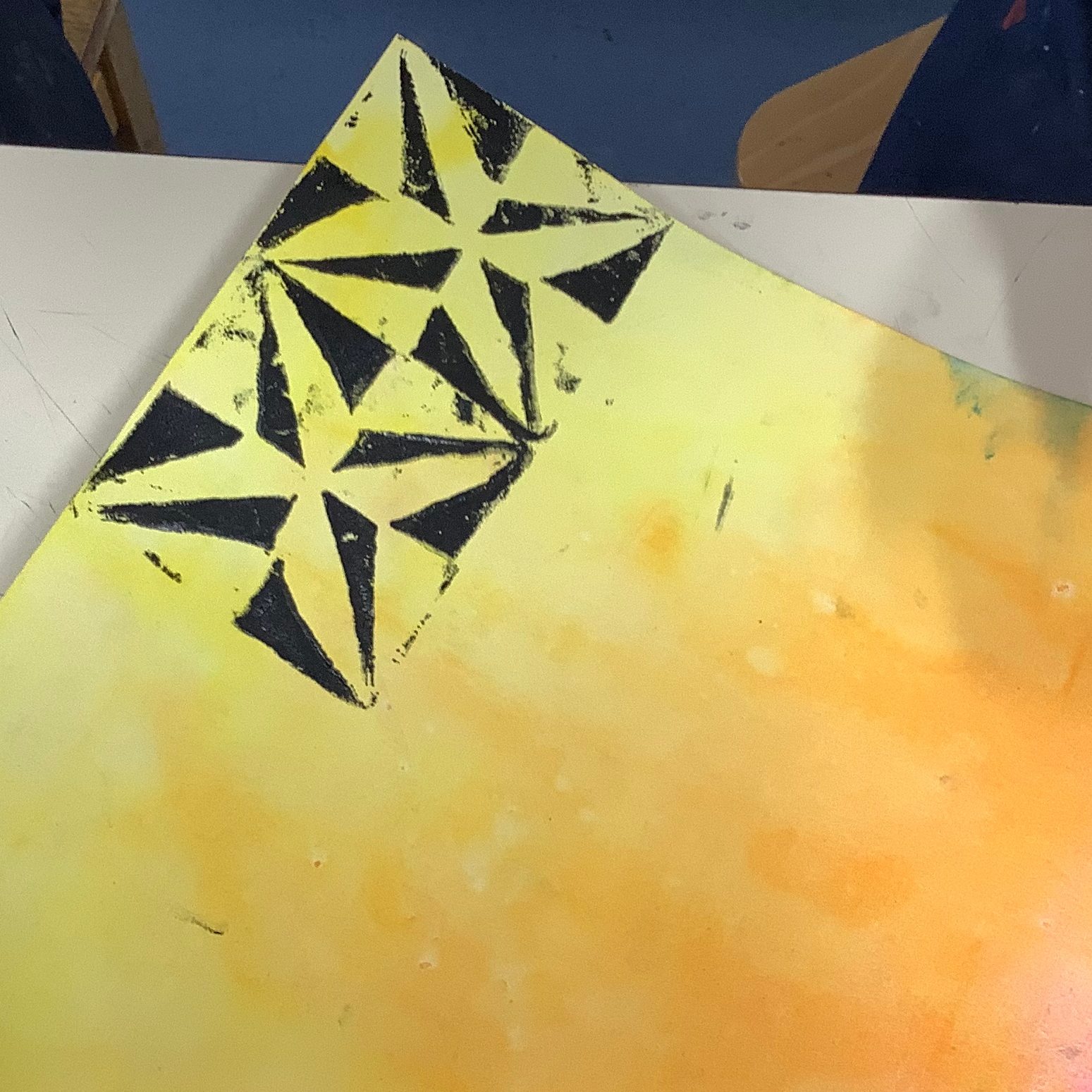 A close-up of a printed design featuring black geometric shapes on a vibrant yellow and orange background, showcasing the texture and color blending in printmaking.