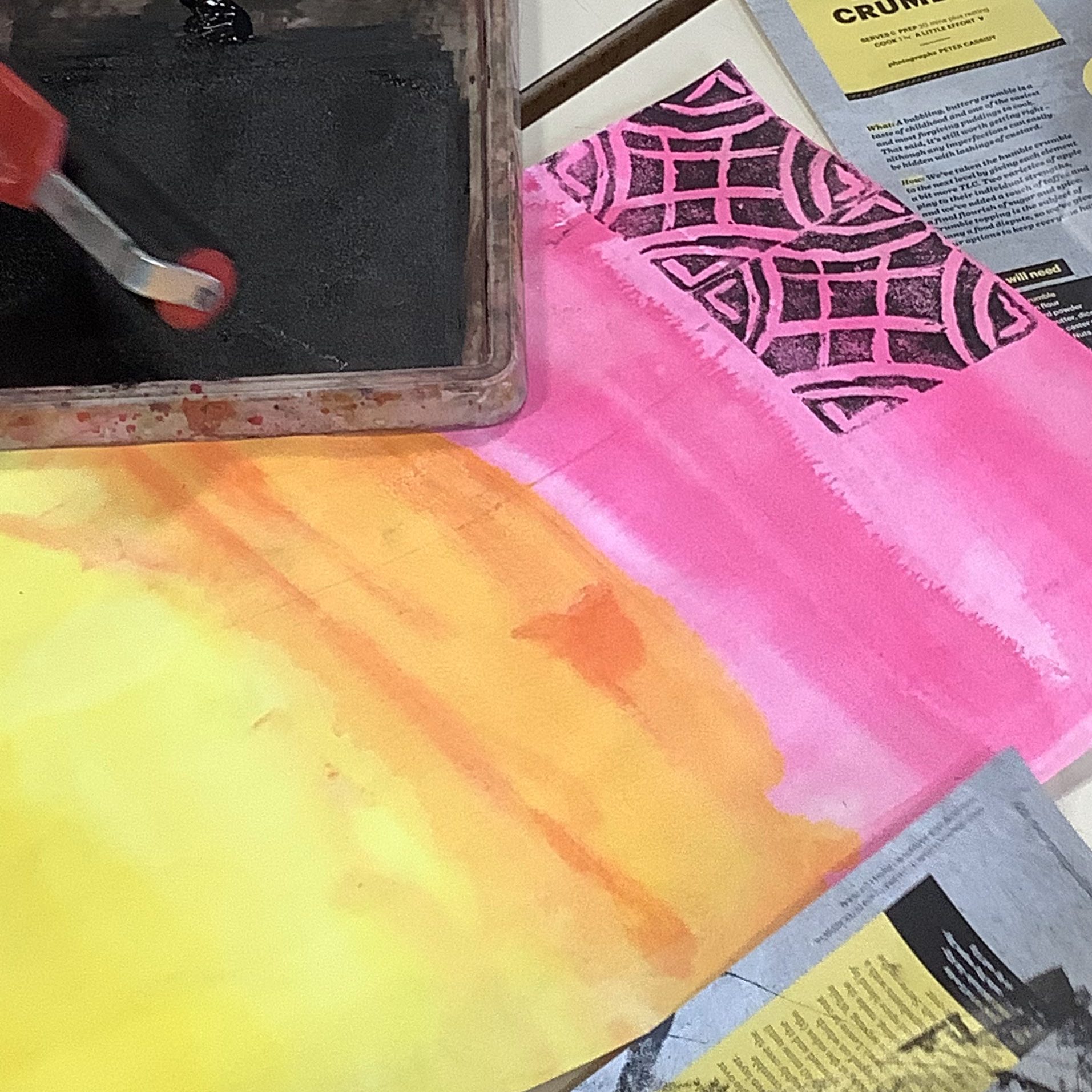 Close-up of a roller applying black paint from a tray onto a vibrant background with yellow, orange, and pink watercolor patterns, surrounded by design papers.