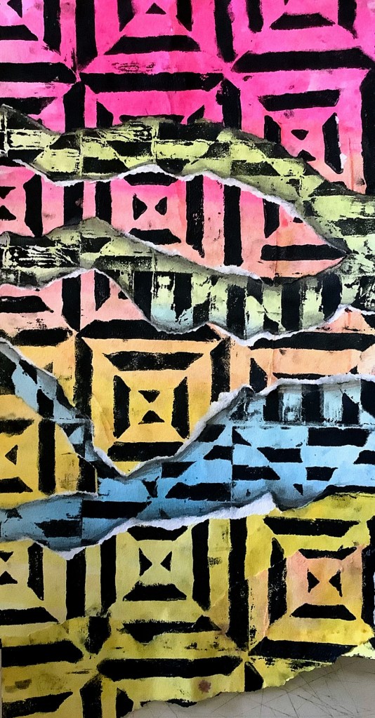 A vibrant printmaking project featuring a colorful pattern of squares in pink, yellow, and blue, with a torn section revealing black geometrical designs underneath.