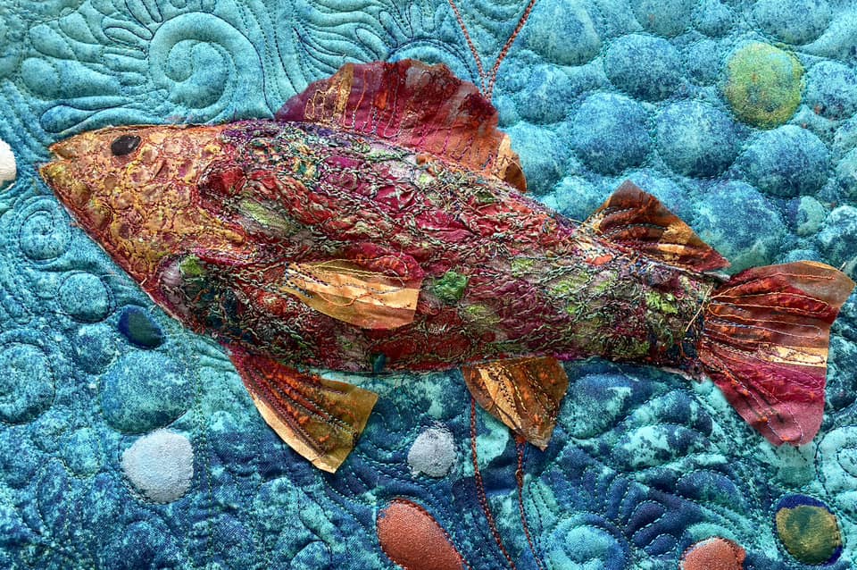 Michelle Mischkulnig - A vibrant textile artwork depicting a fish, intricately crafted with colorful fabrics and textured stitching, set against a backdrop of blue hues and circular patterns.