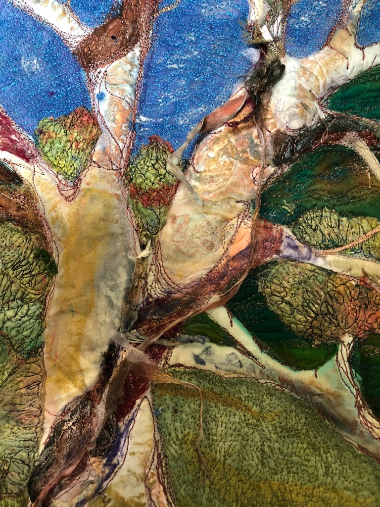 A close-up view of a textile art piece featuring layered fabrics and intricate stitching, depicting a tree with colorful foliage and a blue sky background. Michelle Mischkulnig