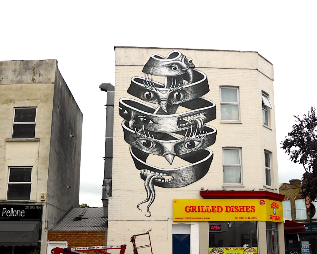 A large-scale mural by street artist Phlegm featuring surreal, interlocking shapes with eyes and tentacle-like forms against the backdrop of an urban building, surrounded by a shop and other structures.