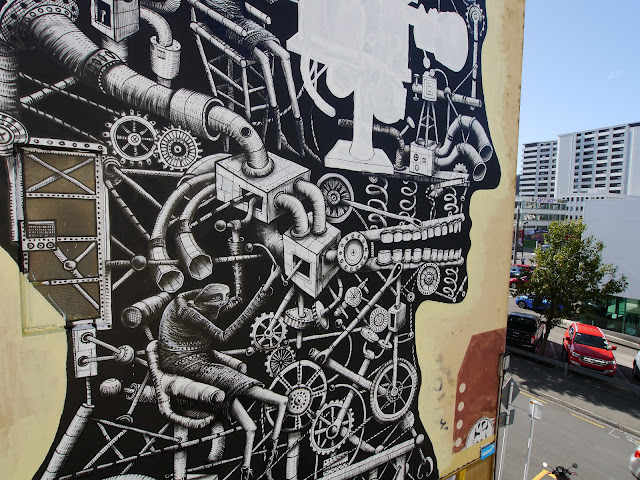 A large-scale black and white mural depicting a stylized human head made of mechanical elements, pipes, and gears, with a figure working within the design.