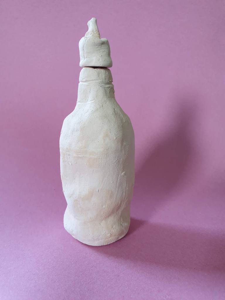 A hand-crafted clay bottle or jar with a rounded body and a narrow neck, displayed against a pink background.