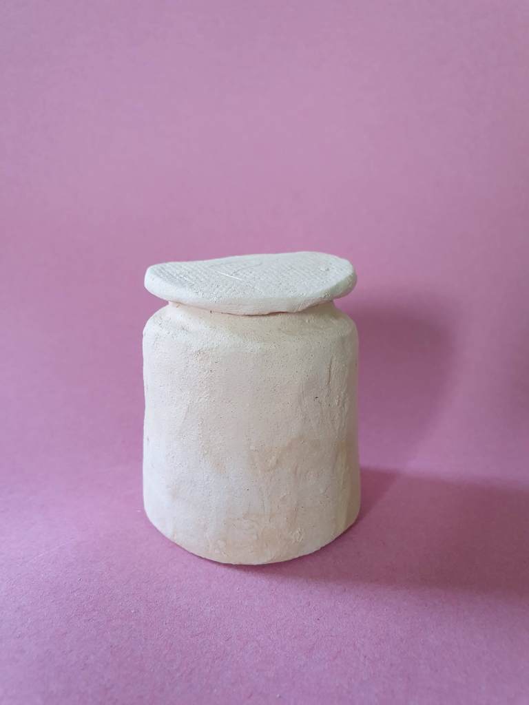 A simple clay jar or bottle with a rounded body and a flat lid, set against a pink background.