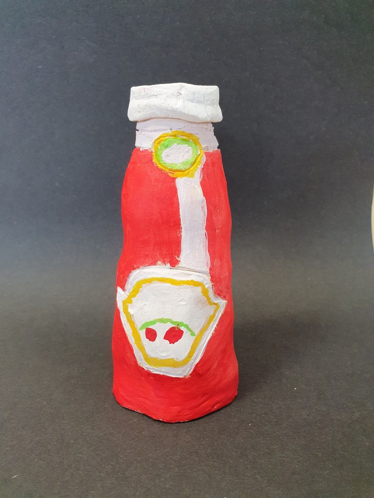 A handmade ceramic ketchup bottle painted in red with a white label and green accents, sitting against a dark background.