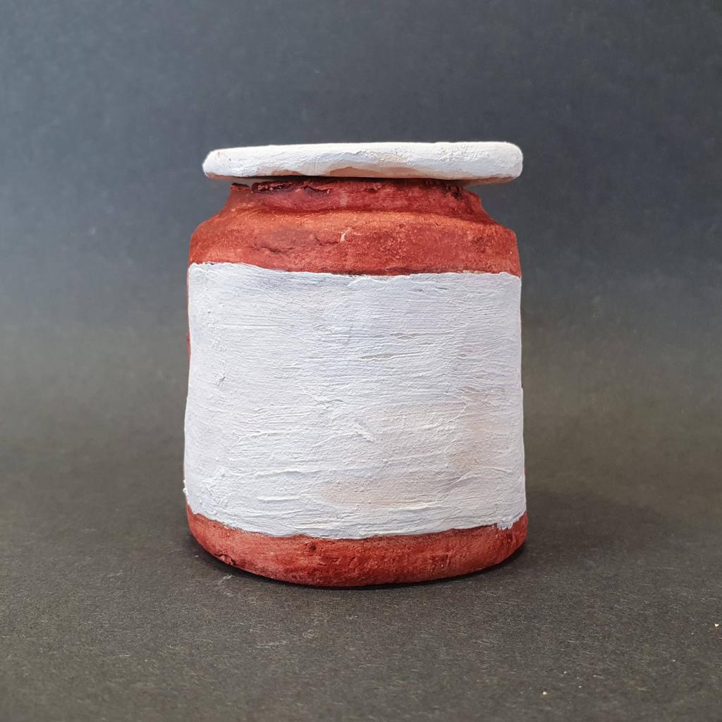 A hand-crafted clay jar with a red base and a white painted label, set against a dark background.