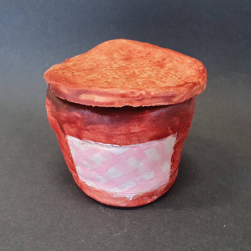 A handmade clay jar or pot with a pink checkered label, resembling a jam jar, set against a black background.