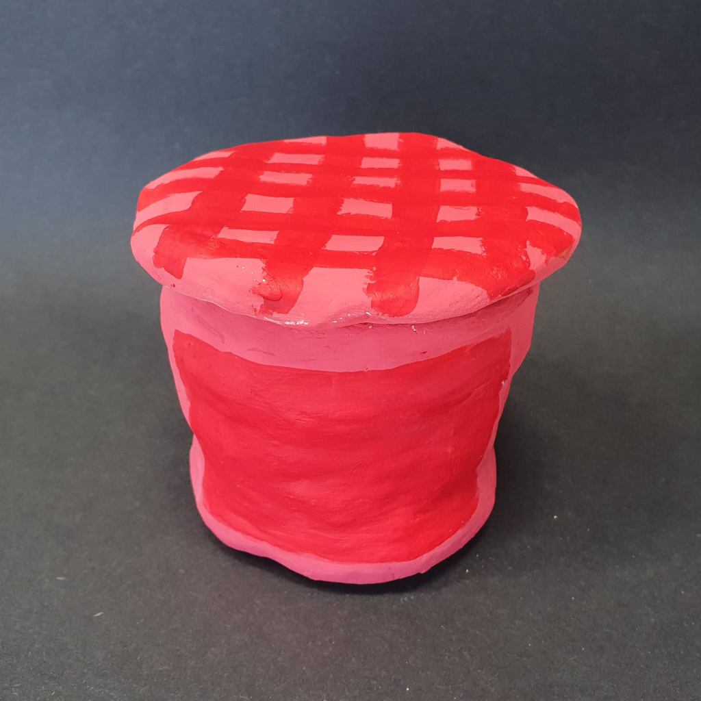 A handcrafted clay jar painted in red with a crisscross pattern on the lid, set against a dark background.