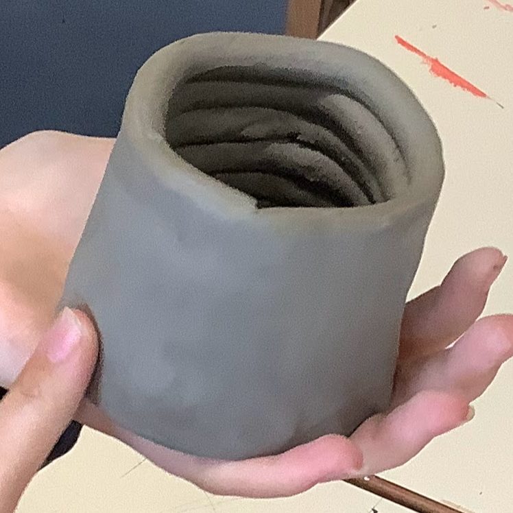 A student holds a hand-built clay jar or container with a coiled design, showcasing the texture and shape of the piece.