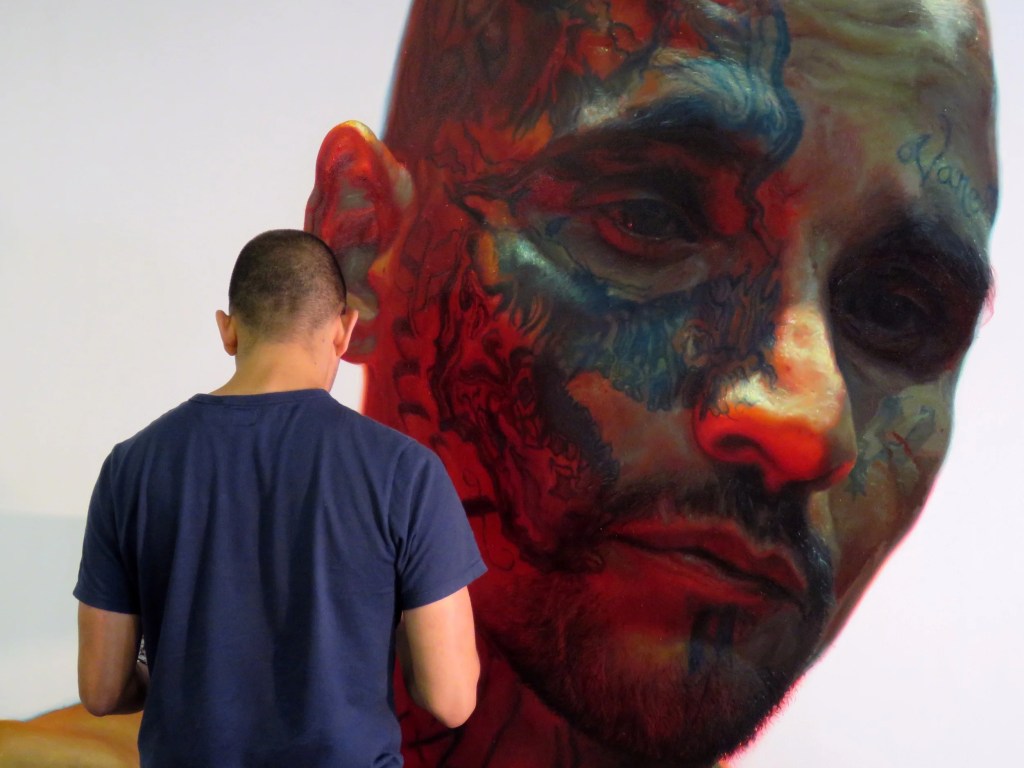 Artist Vincent Valdez is standing with his back to the viewer, focusing on a large, detailed portrait of a tattooed face painted on a wall.
