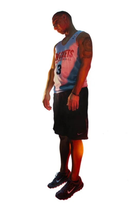 A male basketball player standing with a slight tilt, wearing a blue Rockets jersey and black shorts, showcasing a tattoo on his left arm and black sneakers.