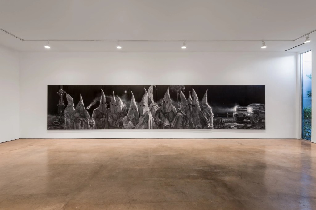 A large monochromatic mural depicting figures in hooded cloaks, arranged in a row, with a pickup truck partially visible on the right side, set against a white gallery wall.