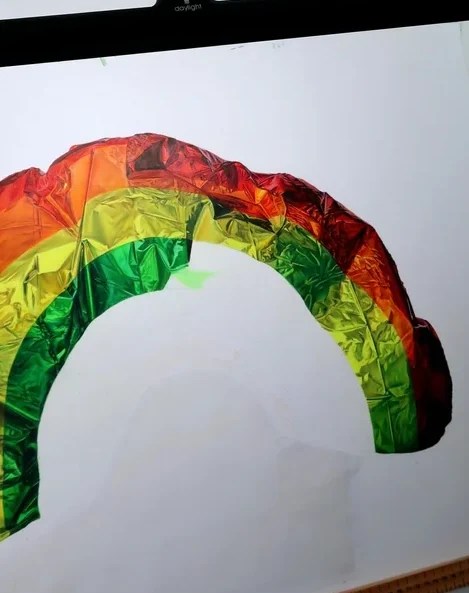 Deflated rainbow-colored inflatable foil balloon with visible creases on a white surface. Coloured pencil artwork